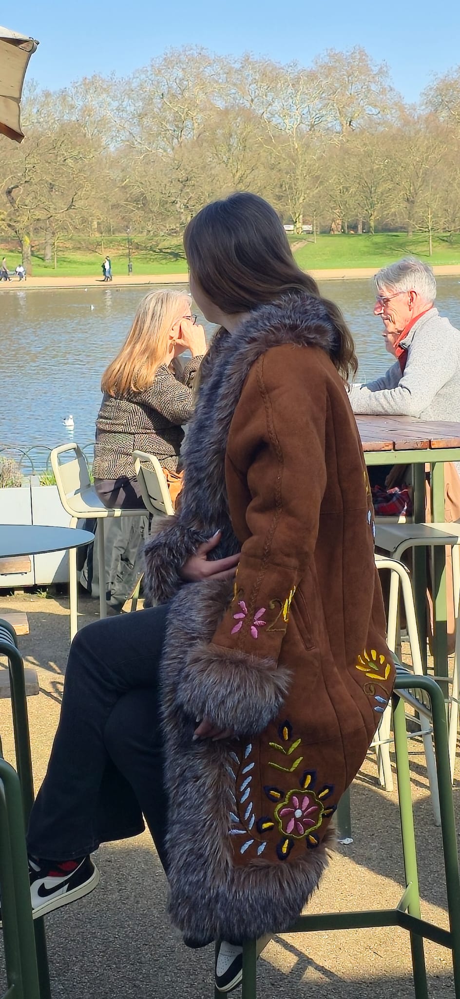 A woman wearing a brown shearling-style coat with gray fur trim and colorful embroidered details sits on a green outdoor patio chair near a lake, with other people and trees in the background.