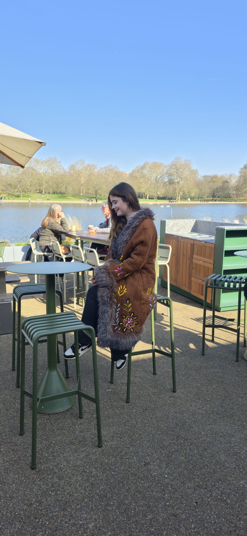 A woman wearing a brown fur-trimmed coat sits at an outdoor patio table next to a lake, with green metal stools and tables around her and trees in the background under a clear blue sky.