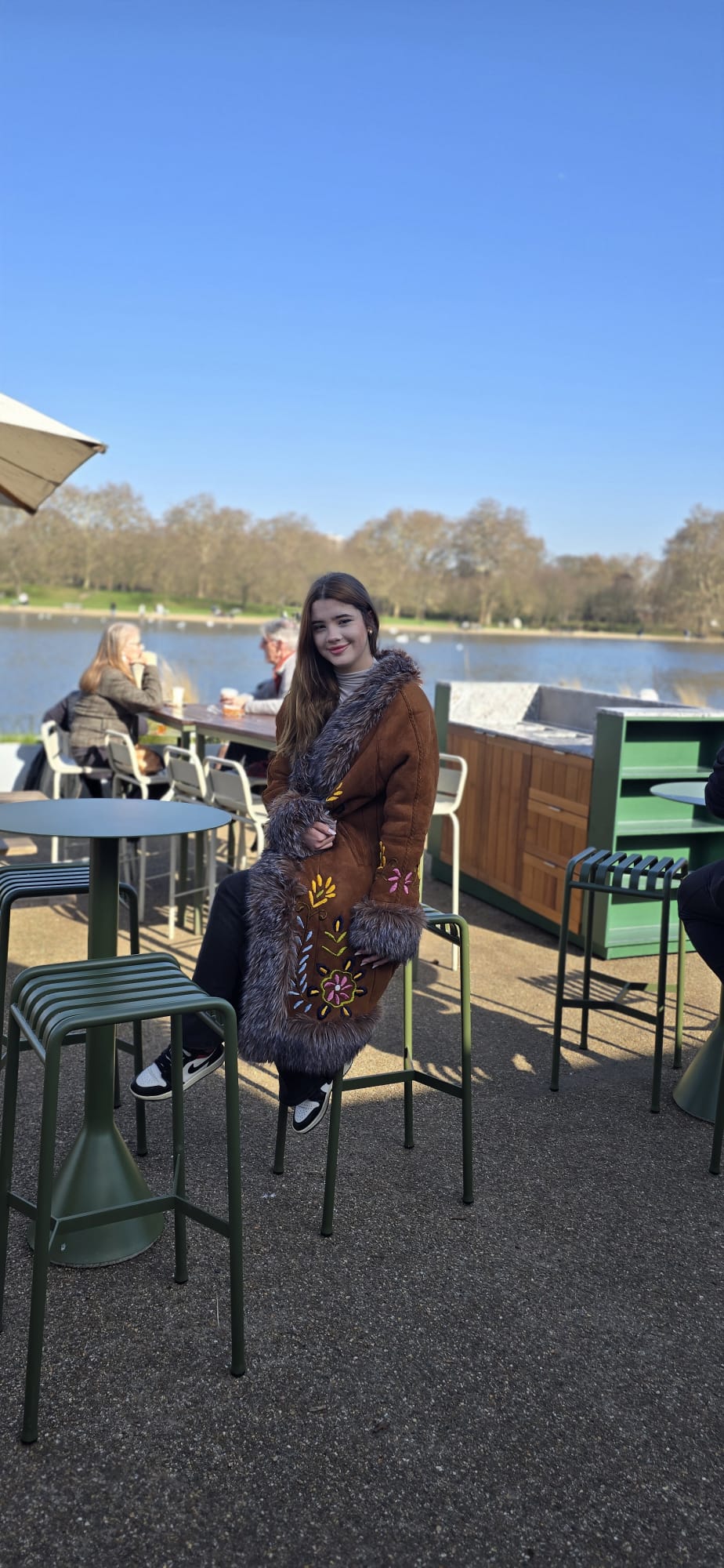 A woman wearing a brown fur-trimmed coat with gold embroidery sits at an outdoor patio table next to a river under a clear blue sky.