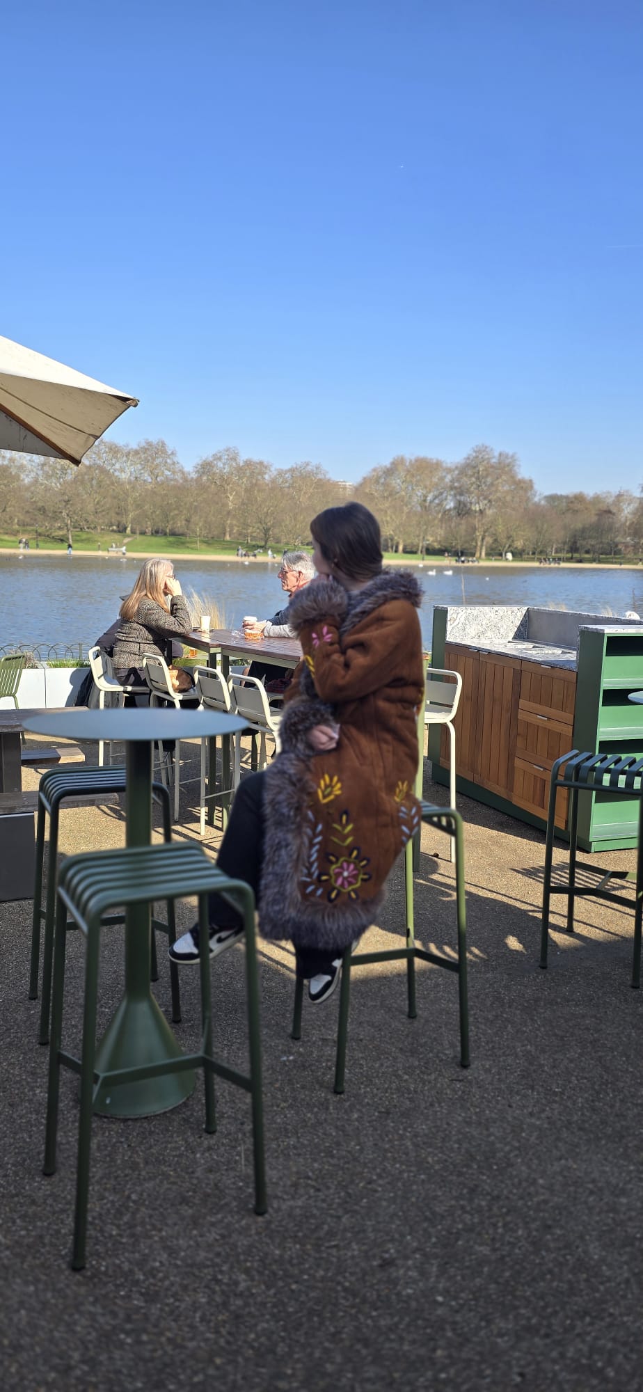 A woman wearing a brown fur-trimmed coat sits on a green metal stool at an outdoor patio table next to a lake, with trees and a blue sky in the background.