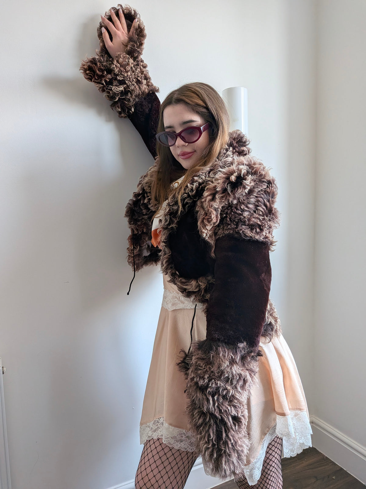 Woman wearing a brown and beige shaggy curly fur cropped jacket with a peach dress, posing against a white wall
