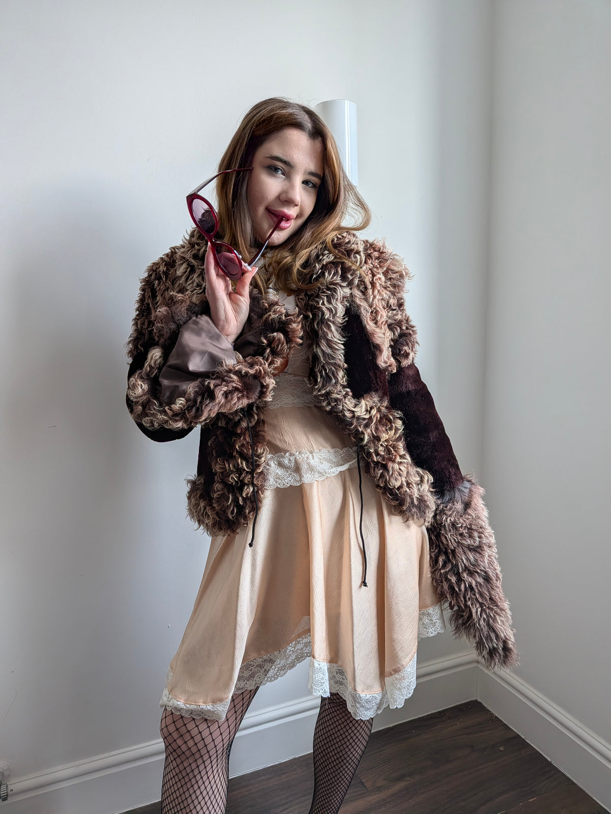Woman posing in a room corner wearing a brown and beige shaggy curly fur jacket over a beige satin dress with lace trim