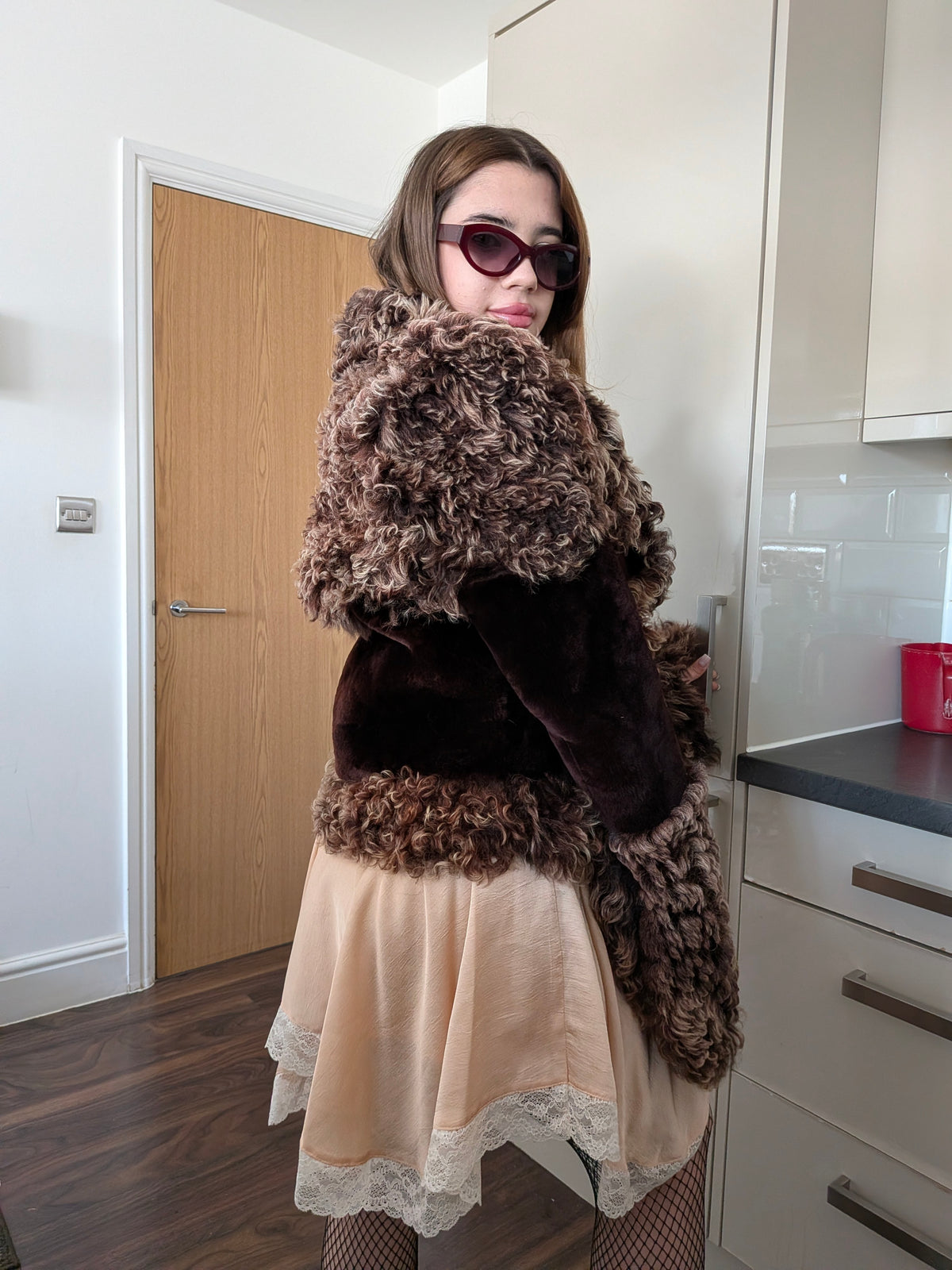 Woman wearing a brown curly fur jacket and a beige skirt with lace trim, standing in a home interior setting