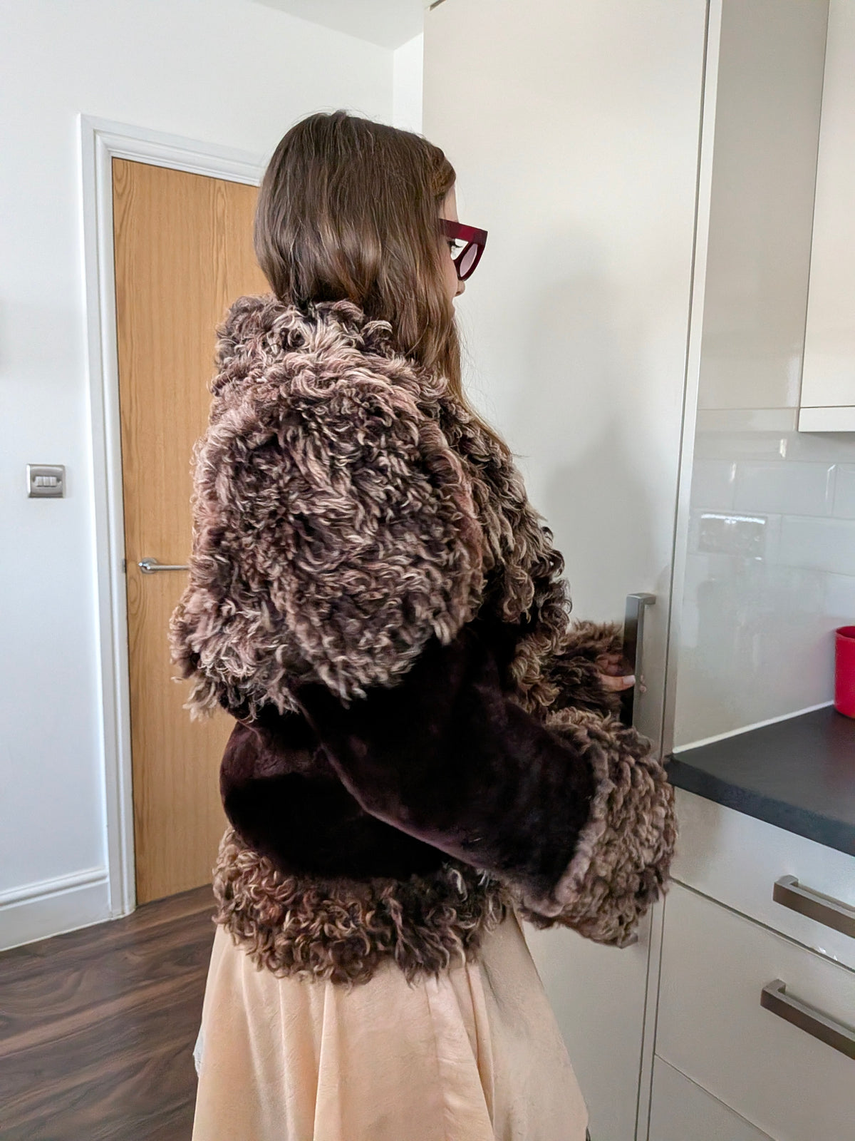 Woman wearing a brown curly fur jacket with dark burgundy velvet sleeves, photographed from behind in a kitchen setting