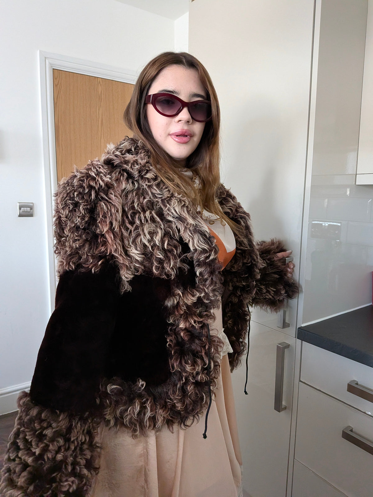 Woman wearing a chunky curly brown and beige fur jacket with dark brown suede panels, standing in a kitchen