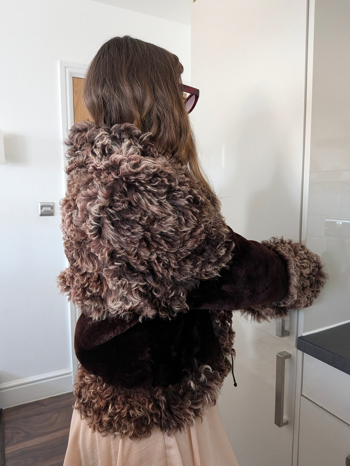 Woman viewed from behind wearing a brown curly fur jacket with dark burgundy velvet sleeves and a matching skirt in a bright indoor setting