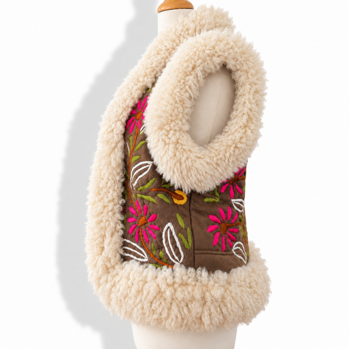 A sleeveless brown vest with a thick cream sherpa lining and a matching large collar, featuring bright pink and green floral embroidery on the front, displayed on a cream mannequin torso against a white background.
