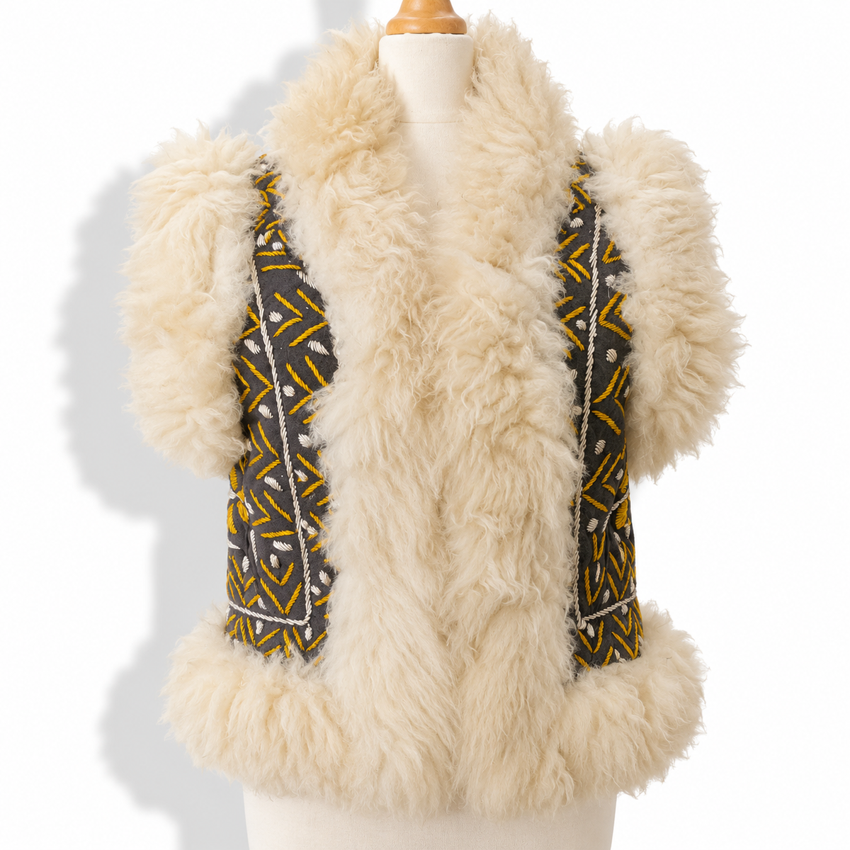 A cream-colored shaggy faux fur vest displayed on a mannequin torso, featuring patterned fabric panels on the front with yellow, black, and white geometric designs.
