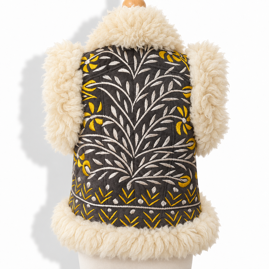 Back view of a hand-embroidered shearling vest with cream faux fur trim, dark grey quilted body, and white and mustard yellow embroidered leaf and floral pattern.