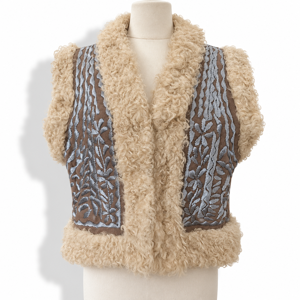 A beige curly faux-fur vest displayed on a cream mannequin torso against a white background. The vest features brown mesh panels on the sides with light blue embroidered floral patterns.