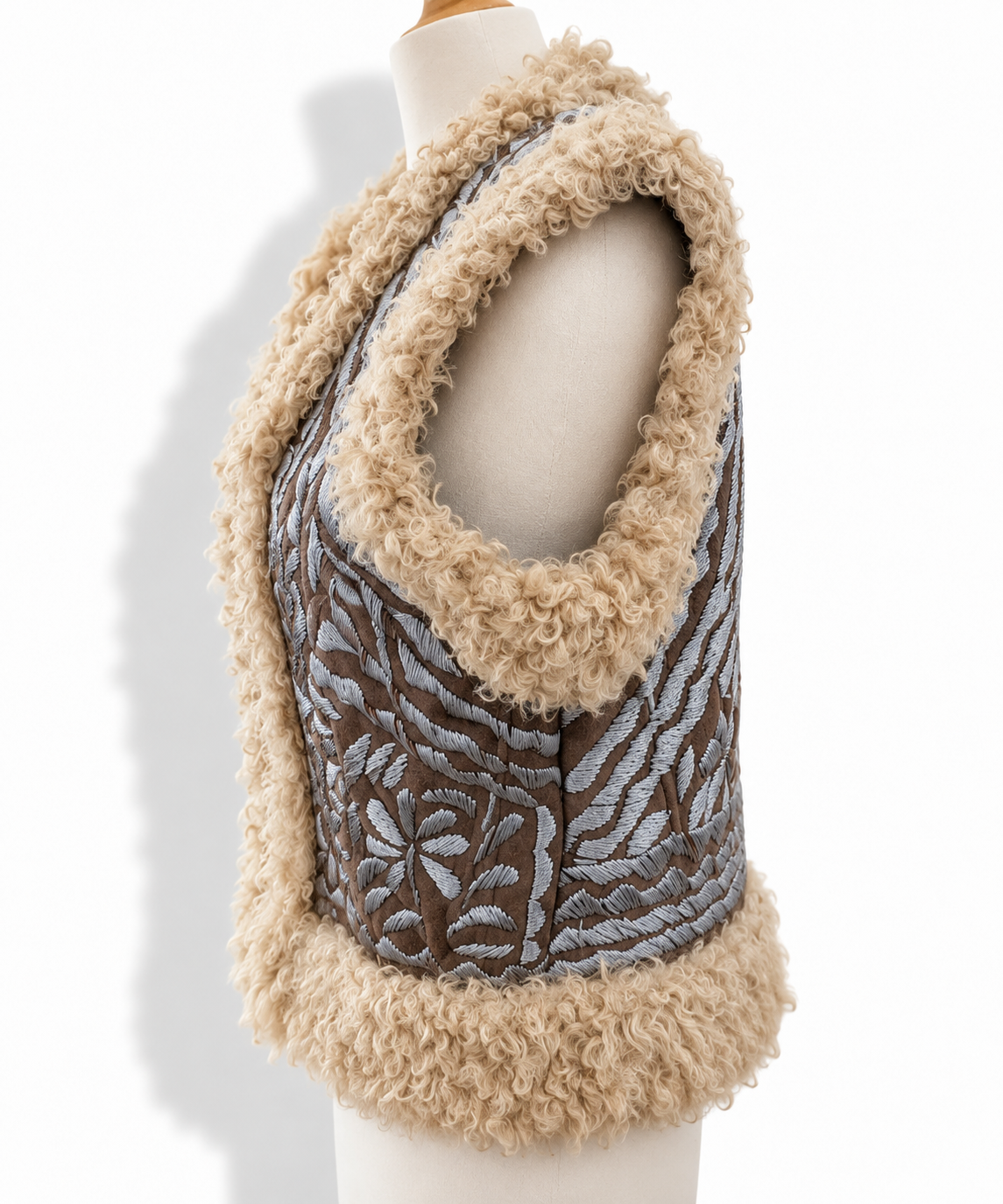 A sleeveless cropped vest displayed on a mannequin torso against a white background. The vest features a brown woven body with a gray floral pattern, trimmed with thick beige curly sherpa fabric.